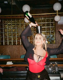 Woman in a red bodysuit and sheer black shrug with braided hair balances a metal shaker on her head while pouring champagne in a retro bar with glass‑block wall, globe lights and barware