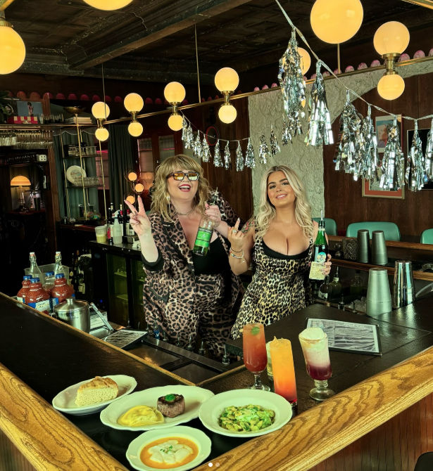 Two bartenders in leopard-print outfits pose behind a retro wooden bar with globe pendant lights and silver tassel decorations, holding liquor bottles; colorful cocktails and plated appetizers on the counter.