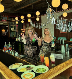Two bartenders in leopard-print outfits pose behind a retro wooden bar with globe pendant lights and silver tassel decorations, holding liquor bottles; colorful cocktails and plated appetizers on the counter.