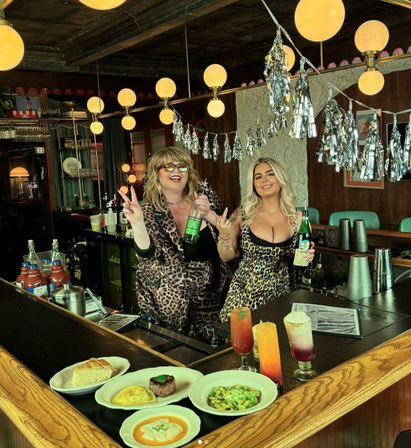 Two bartenders in leopard-print outfits pose behind a retro wooden bar with globe pendant lights and silver tassel decorations, holding liquor bottles; colorful cocktails and plated appetizers on the counter.