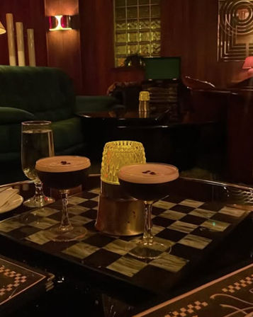 Two espresso martinis topped with coffee-bean garnish on a black-and-white checkered table in a dim, retro-style cocktail lounge with green velvet sofa, amber candlelight and wood-paneled walls.