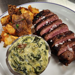 Steakhouse-style sliced medium-rare grilled steak with flaky sea salt, metal ramekin of creamy spinach, and crispy smashed potatoes with chives on a white plate