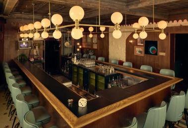 Cozy retro mid-century modern U-shaped bar interior with wood-paneled walls, mint-green swivel stools, black countertop, brass multi-globe pendant lights and a stocked center service area