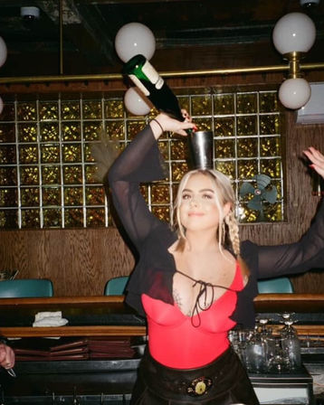 Bartender in a red bodysuit balancing a metal cup on her head while pouring champagne in a retro bar with glass-block wall and globe pendant lights