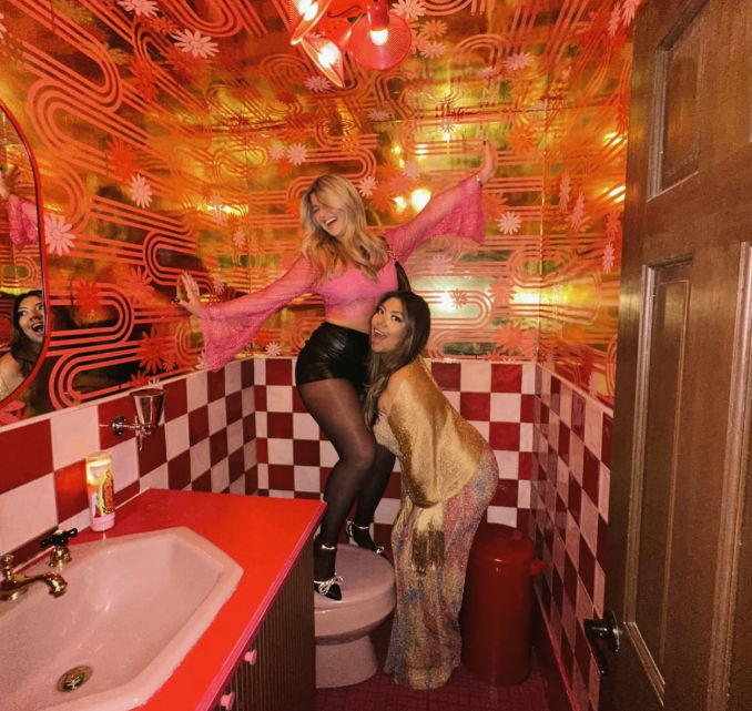 Two friends posing playfully in a retro disco‑themed pink and gold restroom with checkerboard tiles, pink sink and metallic floral wallpaper — one standing on the toilet, the other laughing.