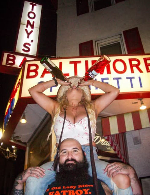 Woman on a man's shoulders chugs from two liquor bottles beneath a glowing neon bar marquee on a lively city street at night.