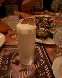 Frothy tall cocktail in a highball glass topped with a dried lemon wheel, sitting on a busy restaurant table with water glasses and shared plates.