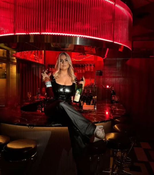 Blonde woman in black leather outfit sitting on a marble bar counter under moody red neon lights, holding two bottles of liquor and striking a confident pose in a stylish cocktail bar.