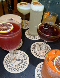 Vibrant assortment of craft cocktails with citrus garnishes and foam, arranged on Madrid-themed coasters at a bar.