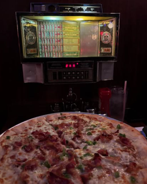 Large pepperoni and green-pepper pizza on a table under a glowing vintage tabletop jukebox selector in a dim retro American diner, with a glass of water and ketchup bottle at the side.
