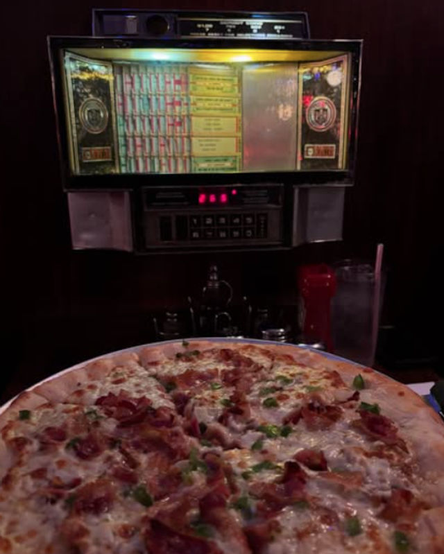 Large pepperoni and green-pepper pizza on a table under a glowing vintage tabletop jukebox selector in a dim retro American diner, with a glass of water and ketchup bottle at the side.