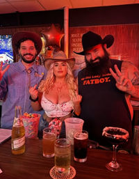 Three people in cowboy hats pose at a red-lit country-themed bar: two men flashing peace signs and a woman in a white lace crop top holding up both middle fingers, with cocktails, a beer bottle, candy jar and a star balloon on the wooden bar.