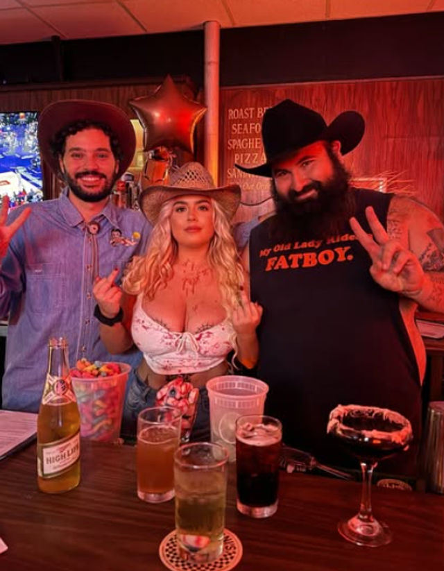 Three people in cowboy hats pose at a red-lit country-themed bar: two men flashing peace signs and a woman in a white lace crop top holding up both middle fingers, with cocktails, a beer bottle, candy jar and a star balloon on the wooden bar.