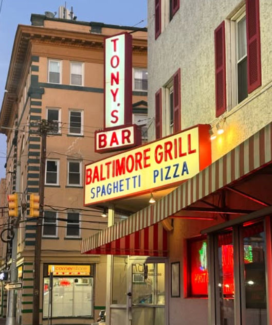 Retro neon marquee for a corner grill and bar on a downtown street advertising spaghetti and pizza above a red-and-white striped awning, framed by multi-story buildings and traffic lights.