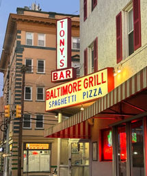 Retro neon marquee for a corner grill and bar on a downtown street advertising spaghetti and pizza above a red-and-white striped awning, framed by multi-story buildings and traffic lights.