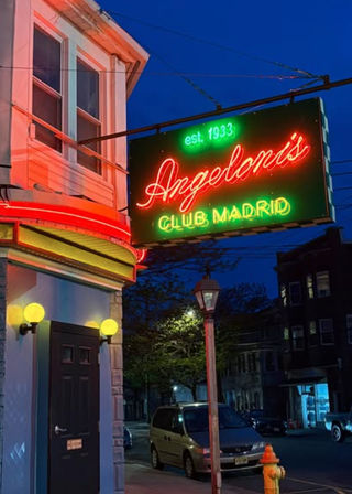 Vintage red-and-green neon sign hangs from a corner building at dusk, glowing curved awning and globe sconces, streetlamp, parked van and yellow fire hydrant on an urban street.