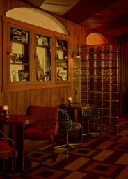 Cozy retro lounge interior with wood-paneled walls, built-in display cabinet, glass block partition, small round tables, patterned floor and warm amber lighting.