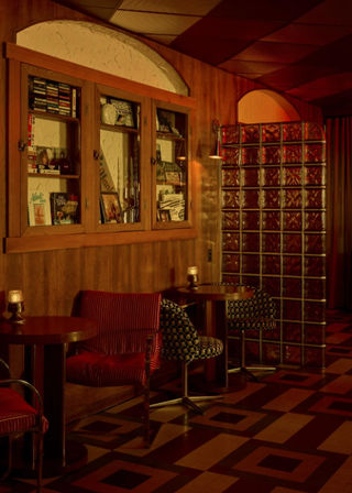 Cozy retro lounge interior with wood-paneled walls, built-in display cabinet, glass block partition, small round tables, patterned floor and warm amber lighting.