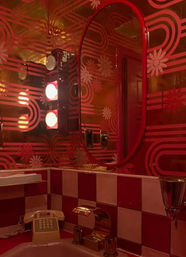 Retro red bathroom with oval mirror, daisy-pattern mirrored wallpaper, checkerboard red-and-white tiles, gold faucet, vintage telephone and glowing vanity lights.
