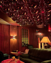 Cozy retro bar lounge interior with wood-paneled walls, plush green sofa, table set with drinks and napkins, warm lamp glow and a mirrored chrome-orb ceiling installation under red ambient lighting.