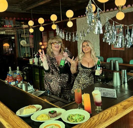 Two bartenders in leopard-print outfits posing behind a retro bar holding bottles, with colorful cocktails and plated brunch dishes on the counter, globe pendant lights and silver streamers overhead creating a festive vintage pub vibe.