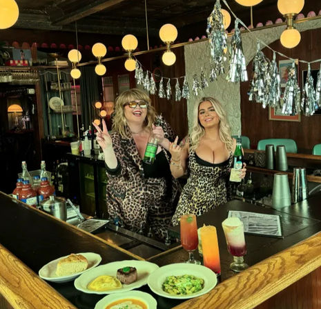 Two bartenders in leopard-print outfits posing behind a retro bar holding bottles, with colorful cocktails and plated brunch dishes on the counter, globe pendant lights and silver streamers overhead creating a festive vintage pub vibe.