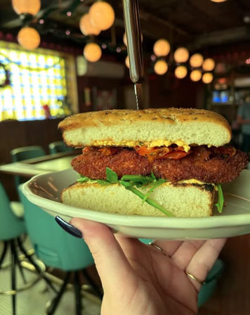 Close-up of a crispy fried chicken sandwich on toasted ciabatta with arugula, roasted tomatoes and sauce, skewered and held on a plate in a retro diner with globe pendant lights and teal barstools.