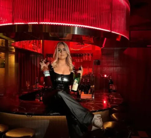 Blonde woman in black leather outfit posing on a marble bar counter holding two liquor bottles under red mood lighting in a nightclub lounge