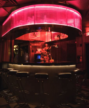 Moody retro cocktail bar interior with a glowing pink circular neon canopy, curved counter lined with black bar stools and a skeleton prop behind the bar