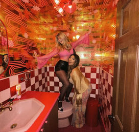 Two friends posing playfully in a neon-lit retro bathroom with orange psychedelic wallpaper, red-and-white checkerboard tiles, and a pink sink and toilet — colorful party photo.