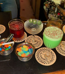Bar countertop with colorful cocktails — deep red drink, martini garnished with three green olives, amber whiskey and a frothy bright green cocktail — plus small bowls of M&M-style chocolates and sour gummy candies.