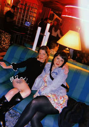 Two women lounging on a blue-striped sofa in a cozy red-lit bar or lounge — one in a black dress and boots with a visible thigh tattoo, the other in a floral dress and denim jacket, lamp glowing and patrons mingling in the background.