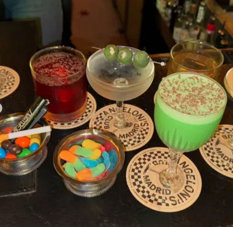 Bar countertop with colorful cocktails — green cream cocktail, olive-topped martini and red drink — plus small bowls of gummy candies in Madrid