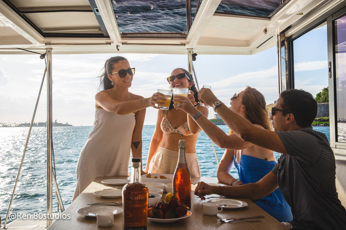 Four friends toasting with drinks on a sunlit yacht deck over blue ocean near a tropical coastline, table set with bottles, fruit and plates — summer boat getaway