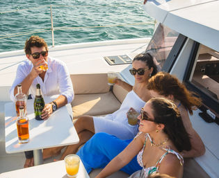 Four friends relaxing on a yacht deck, sipping cocktails and champagne under sunny skies with blue ocean in the background.