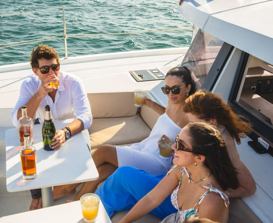 Four friends relaxing on a yacht deck, sipping cocktails and champagne under sunny skies with blue ocean in the background.