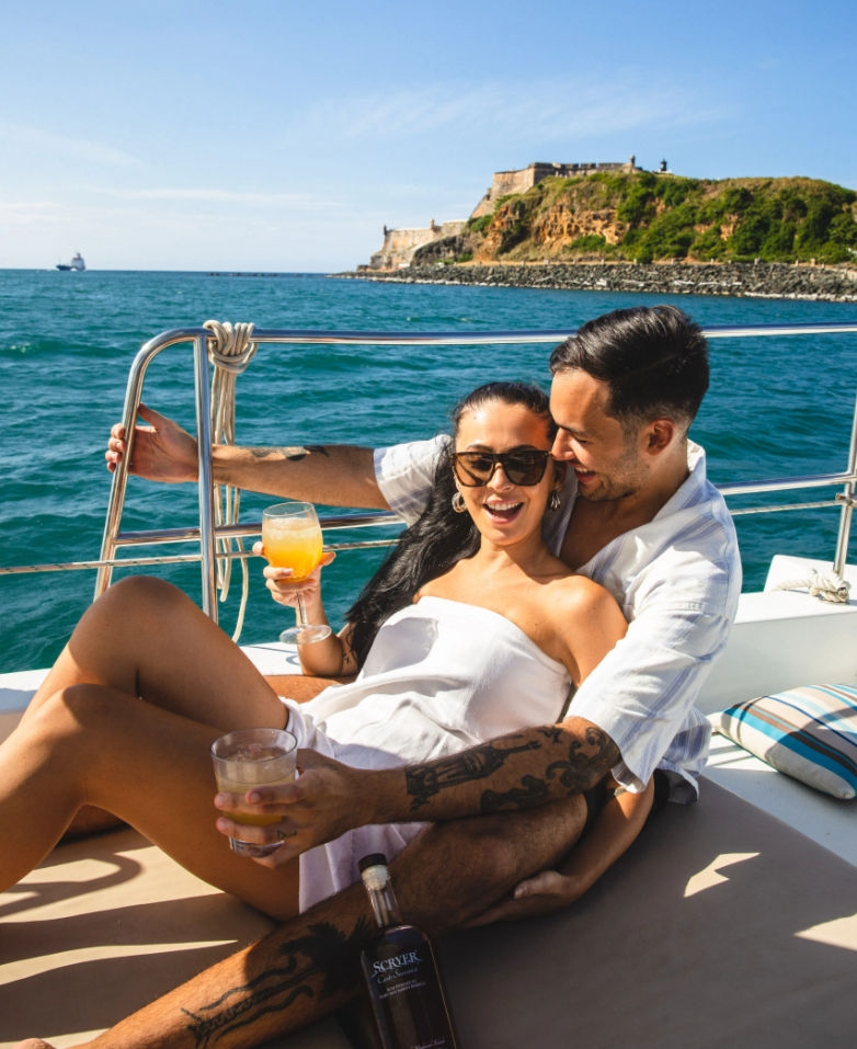 Couple relaxing on a sunny yacht, holding cocktails and laughing with turquoise ocean and a historic cliffside fort on the shoreline behind them.