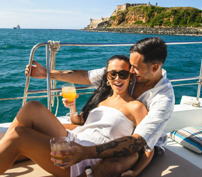 Couple relaxing on a sunny yacht, holding cocktails and laughing with turquoise ocean and a historic cliffside fort on the shoreline behind them.