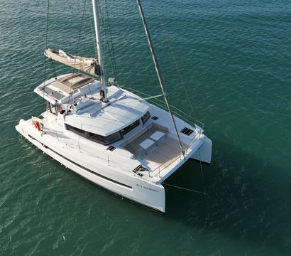 Aerial view of a white luxury sailing catamaran anchored on calm turquoise water, showing twin hulls, a furled mainsail and spacious forward deck seating.