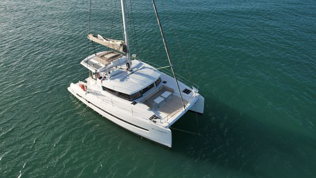 Aerial view of a white luxury sailing catamaran anchored on calm turquoise water, showing twin hulls, a furled mainsail and spacious forward deck seating.