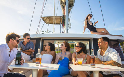 Smiling group of friends enjoying cocktails on a sunlit sailboat deck, chatting while one woman sunbathes on the cabin roof near a marina