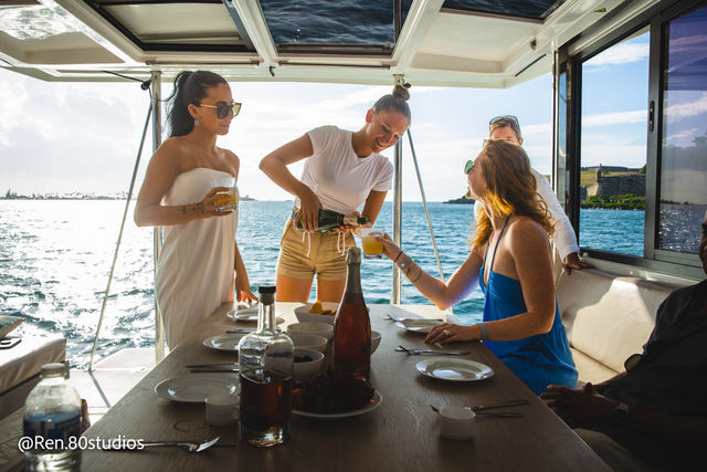 Sunny yacht brunch with people—server pours mimosas at a table set with plates and bottles, open ocean and coastline visible through the cabin