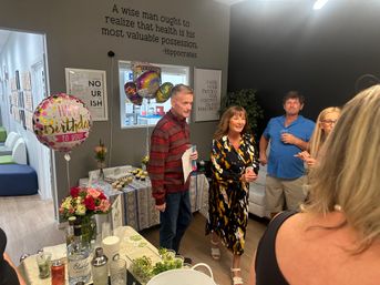 Cheerful indoor birthday celebration in a modern office reception area with 'Happy Birthday' balloons, cupcakes and flowers on a table, and guests chatting beneath a framed health quote on the wall.