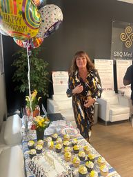 Woman smiling at an indoor med-spa reception birthday celebration with colorful "Happy Birthday" balloons, cake and yellow-topped cupcakes on a decorated table