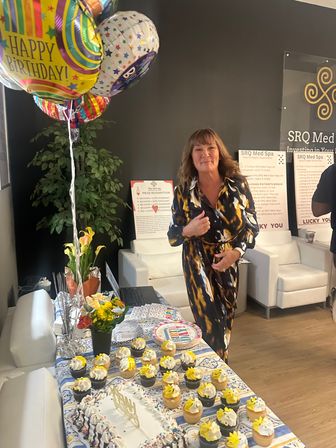 Woman smiling at an indoor med-spa reception birthday celebration with colorful "Happy Birthday" balloons, cake and yellow-topped cupcakes on a decorated table
