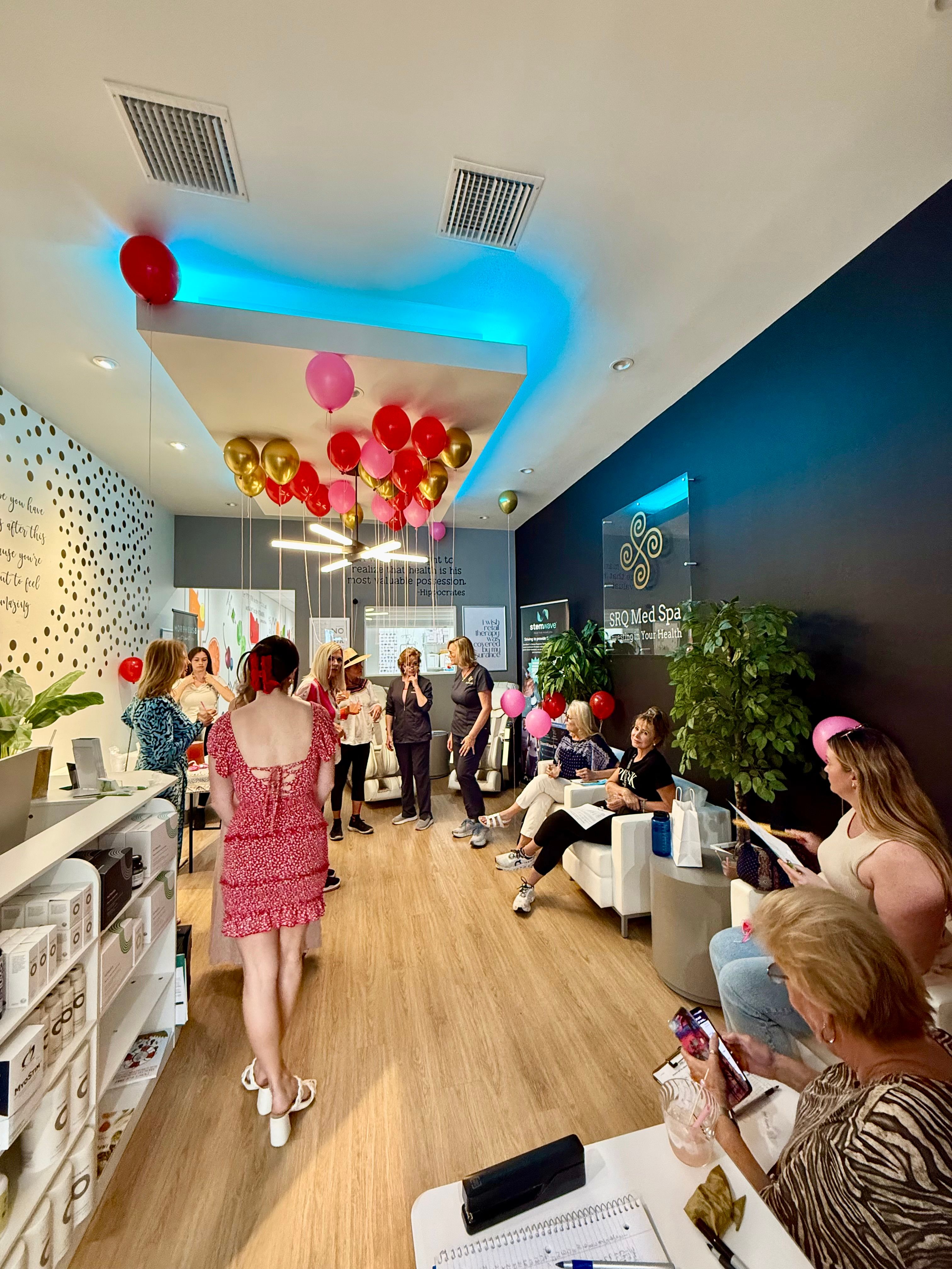 Women at a lively med-spa celebration in a bright waiting room with pink and gold balloons, blue ceiling lighting, white sofas, wood floors and potted plants.