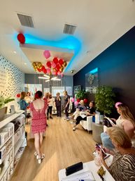 Women at a lively med-spa celebration in a bright waiting room with pink and gold balloons, blue ceiling lighting, white sofas, wood floors and potted plants.