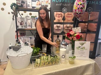 Woman mixing cucumber vodka cocktails at a skincare pop-up table with ice bucket, cucumber skewers, cocktail ingredients, roses, balloon, and before-and-after facial treatment display