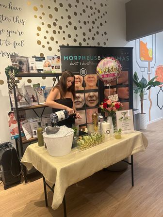 Woman mixing cocktails at a birthday drink station inside a skincare clinic/medspa, table with ice bucket, garnishes and flowers, birthday balloon and skincare before-and-after posters on a gold polka-dot wall.
