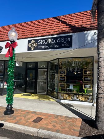 Bright med-spa storefront on a sunny shopping street with a red-tiled awning, ornate gold window lettering reading The Picture of Health, and a lamppost wrapped in green garland and a red bow.